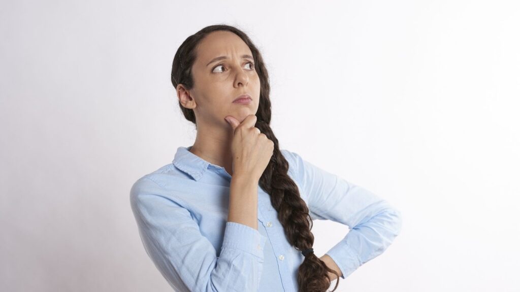 White woman with long brown hair wearing a light blue shirt. She is furrowing her brow and has one hand holding her chin and the other resting on her waist, as if thinking.