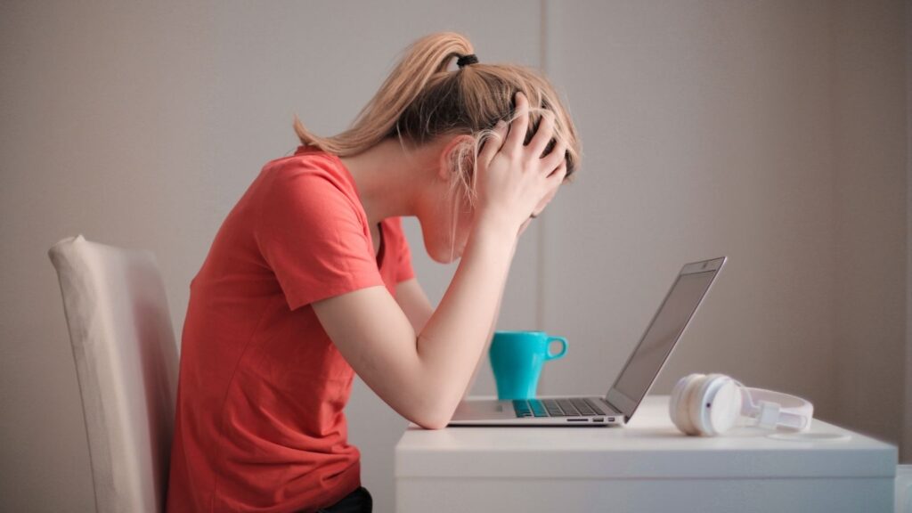 White woman with blonde hair up in a ponytail wearing an orange t-shirt. She's sitting at a desk with her elbows on the table and her hands on her head. She's looking down at a laptop, and the whole thing suggests frustration.