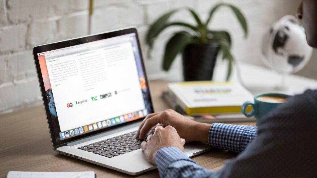 Hands on a laptop keyboard. Laptop is on a desk. On the screen is a webpage.