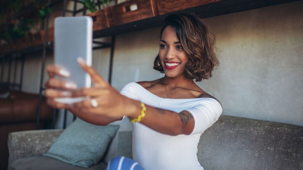 Smiling Black woman holding a cellphone at arms length while sitting in  a modern cafe, She's taking a selfie.