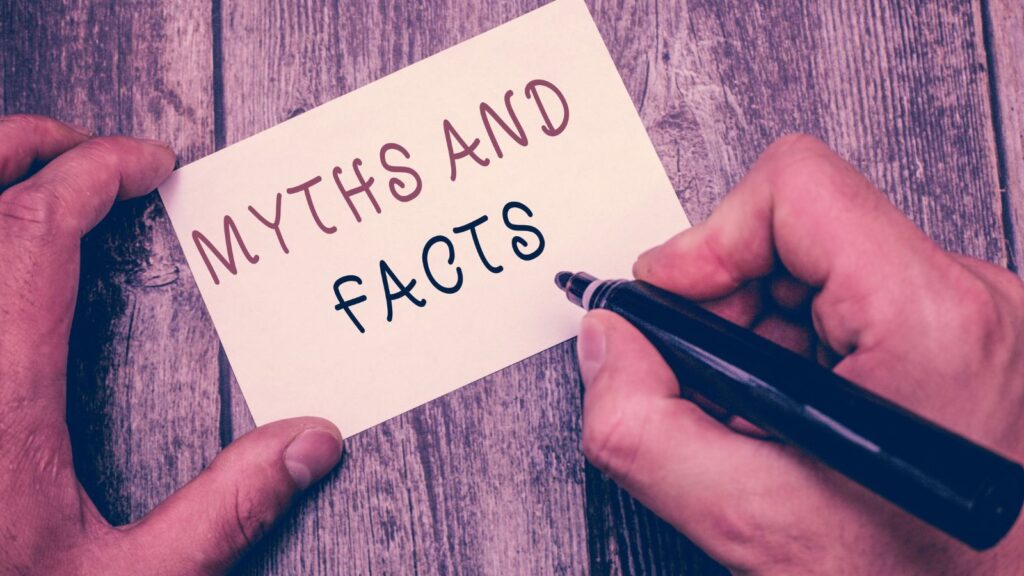 Notecard with text MYTHS AND FACTS. Left hand holding card in place; right hand holding a marker over card. Background is a distressed wooden table or wall.