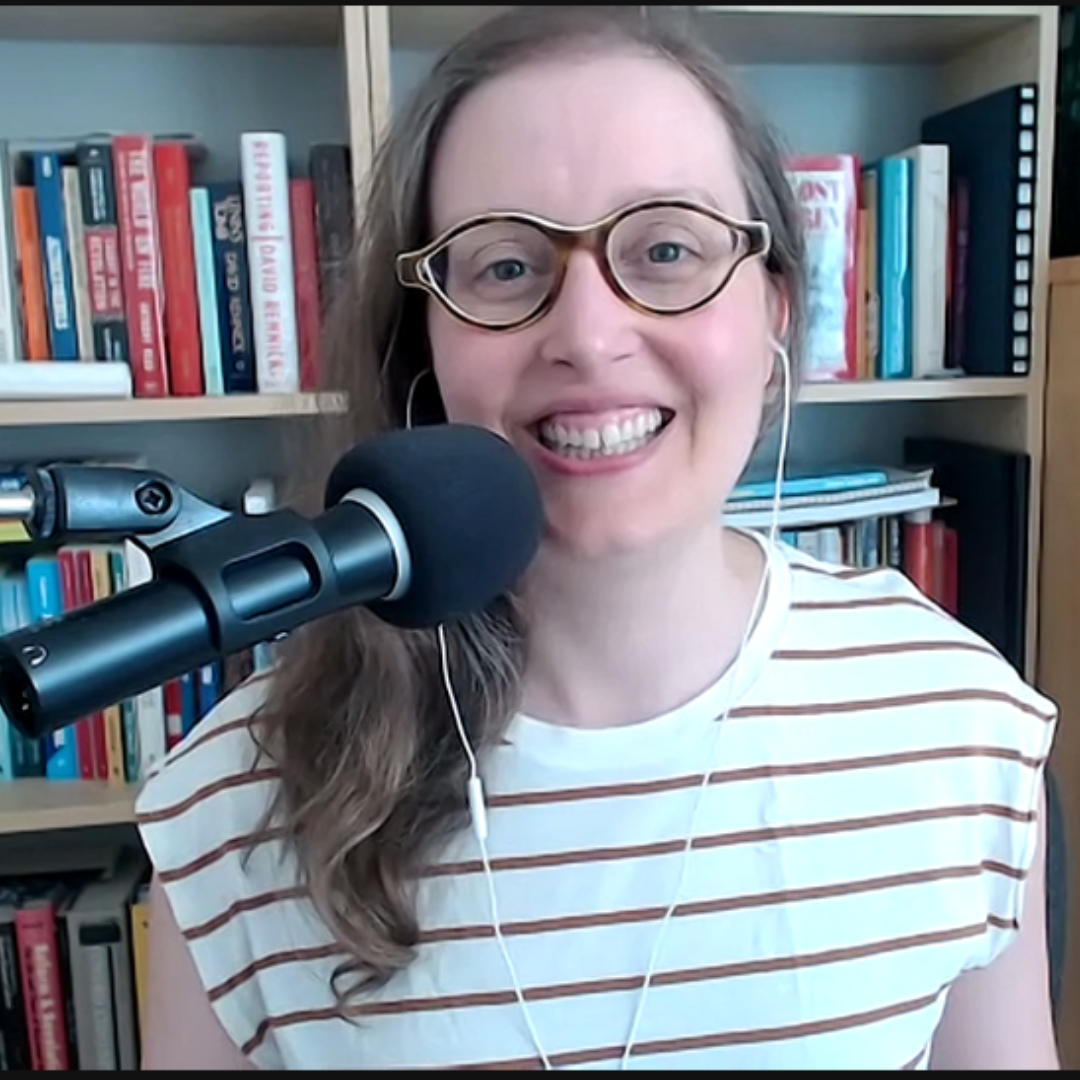 In this image, a person with a joyful smile is seen, ready to engage in a conversation or presentation. They're wearing a white and tan striped shirt and stylish glasses, with earbuds in. A professional microphone is positioned in front of them, indicating they're prepared for clear audio communication. The backdrop features an organized bookshelf filled with a range of books that add a touch of intellectual depth to the setting, alongside a wooden cabinet that complements the cozy, yet professional home office environment.