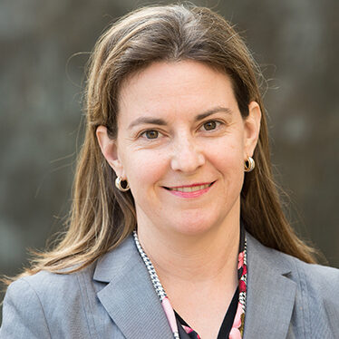 Kenna Barrett, a person with long brown hair smiles at the camera, wearing a gray blazer and floral-patterned blouse, standing against a blurred background.