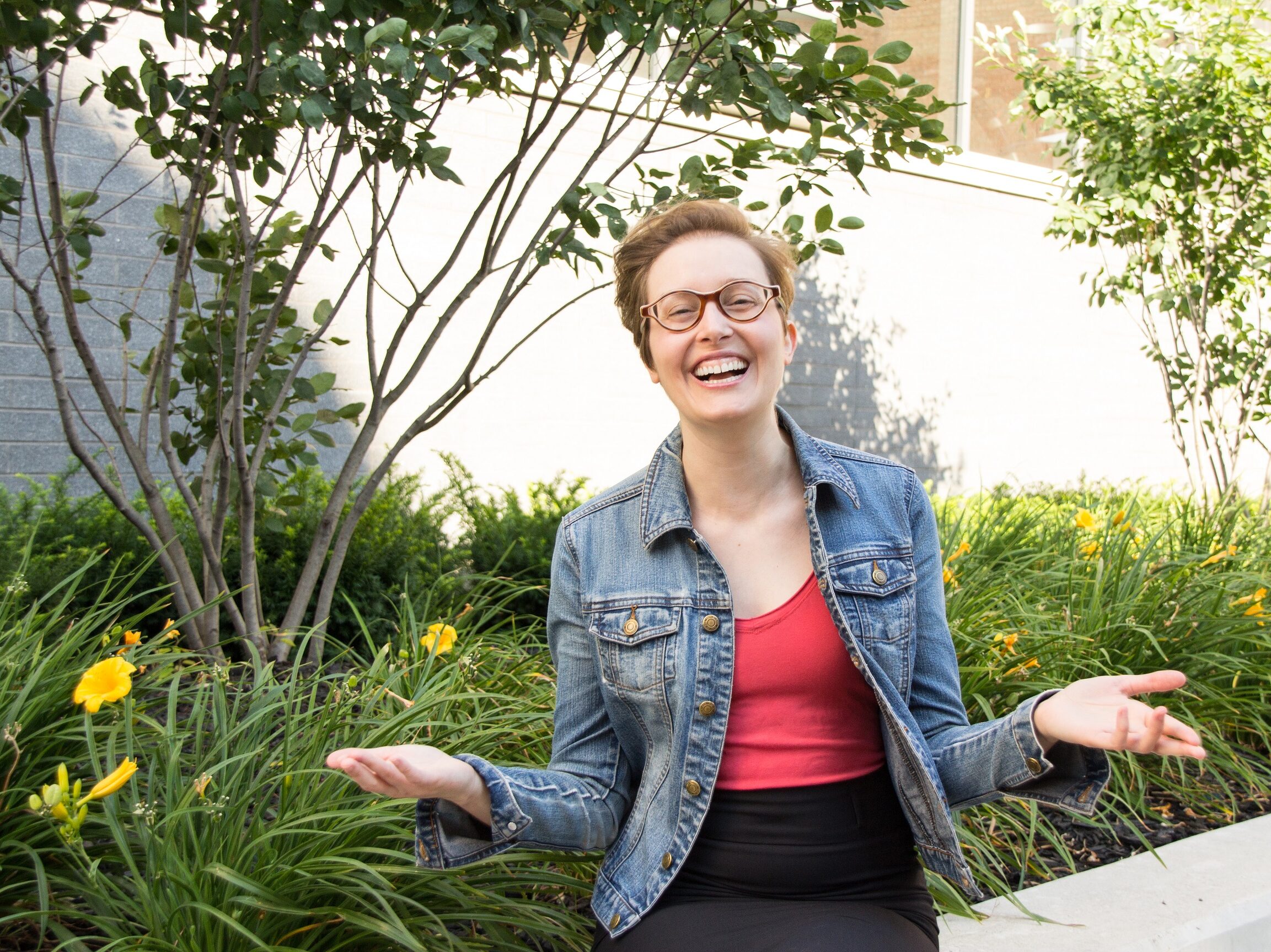 A person with short hair and glasses is sitting outside, smiling with arms outstretched. They are wearing a denim jacket and red top, celebrating their new PhD career. Green plants and yellow flowers surround them, framed by a brick wall in the background.