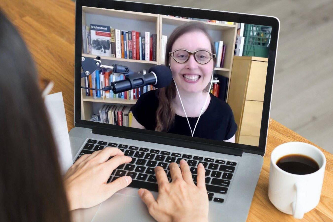 A person participates in a video call on a laptop with a smiling woman wearing glasses and headphones, sitting in front of a bookshelf. A cup of coffee sits on the wooden table beside the laptop.