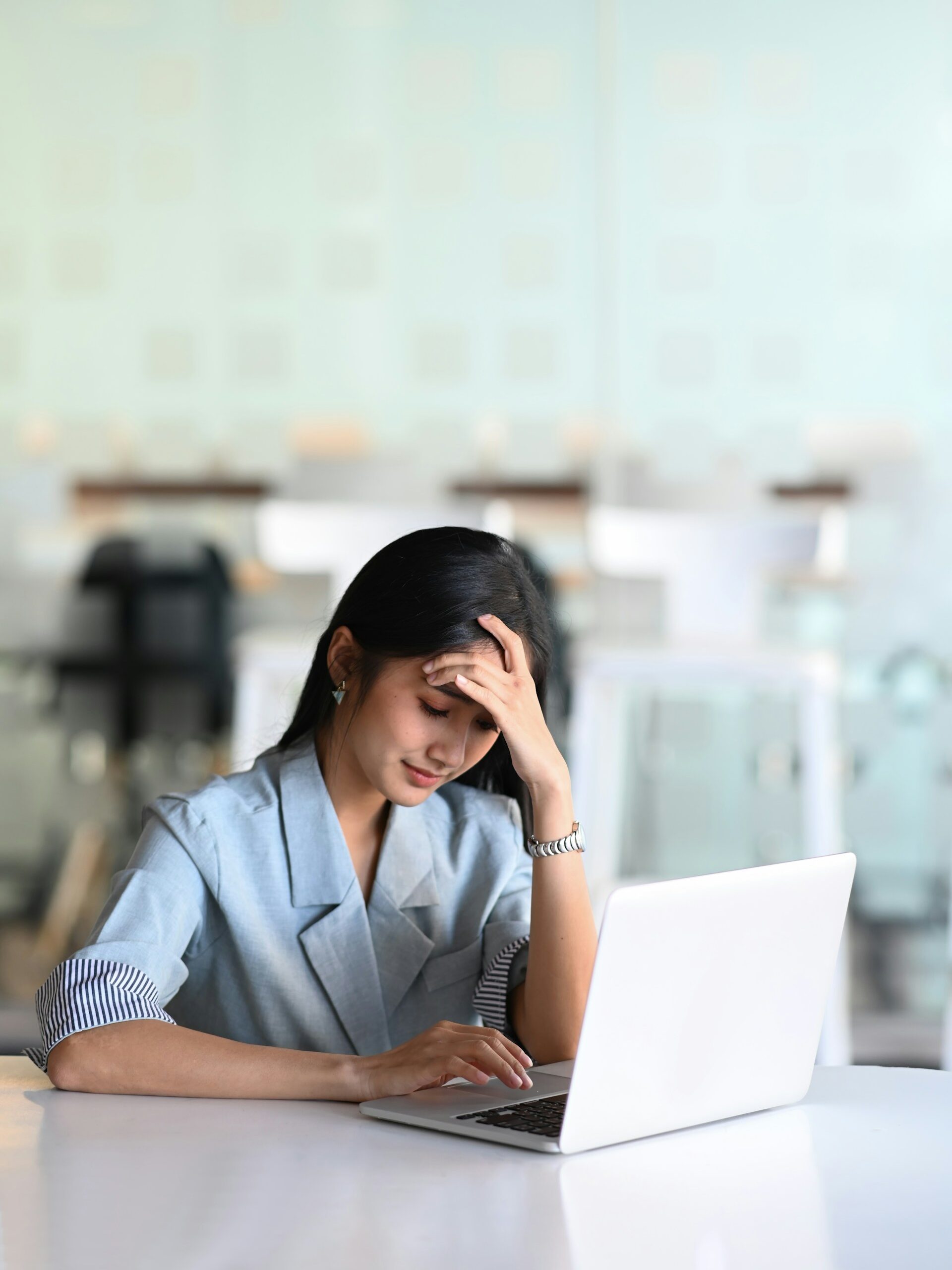 A woman sits at a desk with a laptop, resting her head on one hand and looking stressed or tired. The background shows a blurred, modern office setting with chairs and hanging lights.